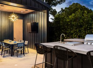 Modern outdoor kitchen and dining area with sleek furniture, grill, and stylish lighting, surrounded by lush greenery.